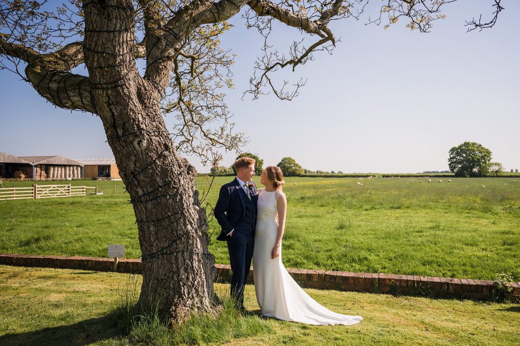 Hornington Manor Wedding - bride and groom posing next to tree with fields behind them