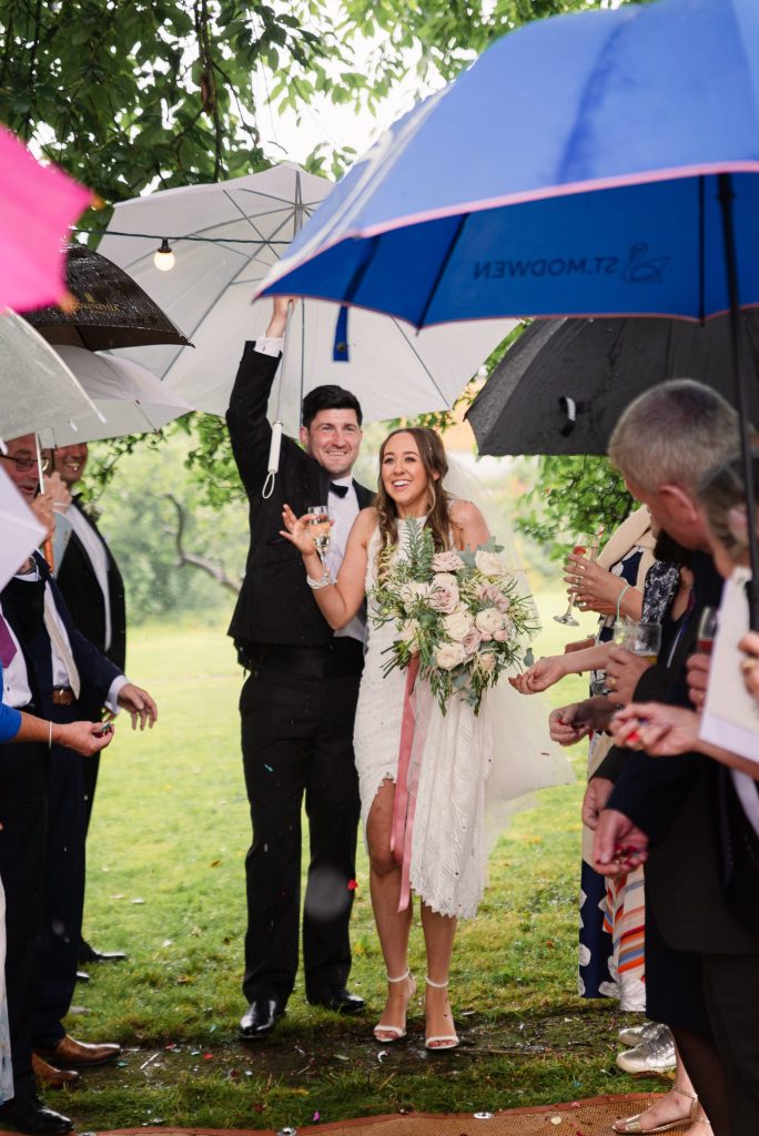 Bride and groom smiling under umbrellas walking through guests at a rainy Tadcaster countryside wedding, Yorkshire.