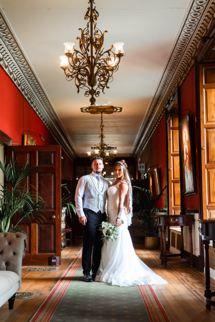 Bride and groom posing under a Victorian chandelier in the corridor of Swinton Park Estate Hotel, luxury Yorkshire wedding.